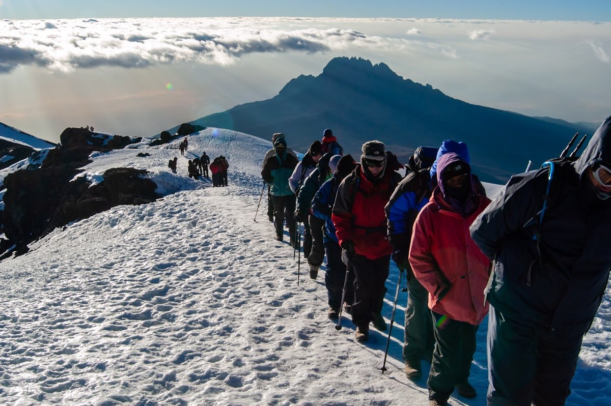 Climbers lined up on the snow-covered crater rim of Kilimanjaro above the cloud layer, with Mawenzi peak in the background
