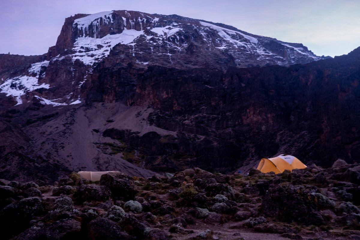 Orange tents pitched in a remote high-altitude camp on Kilimanjaro's rocky northern flank with snow-capped cliffs above, Tanzania